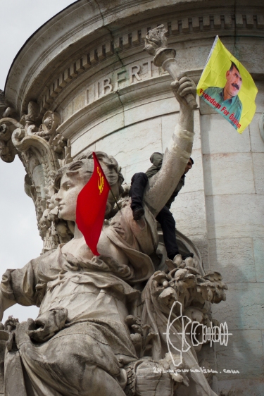 A protestor clims on a statue and attaches a "Freedom for Öcalan" flag below the words "Liberty"
